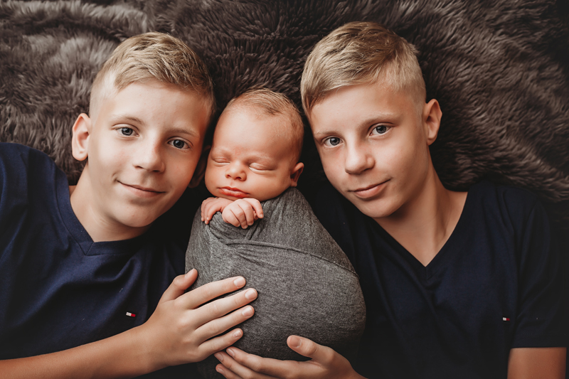 Midlothian, Virginia newborn pics, newborn baby boy swaddled in gray fabric on gray backdrop with twin blond brothers lying beside him