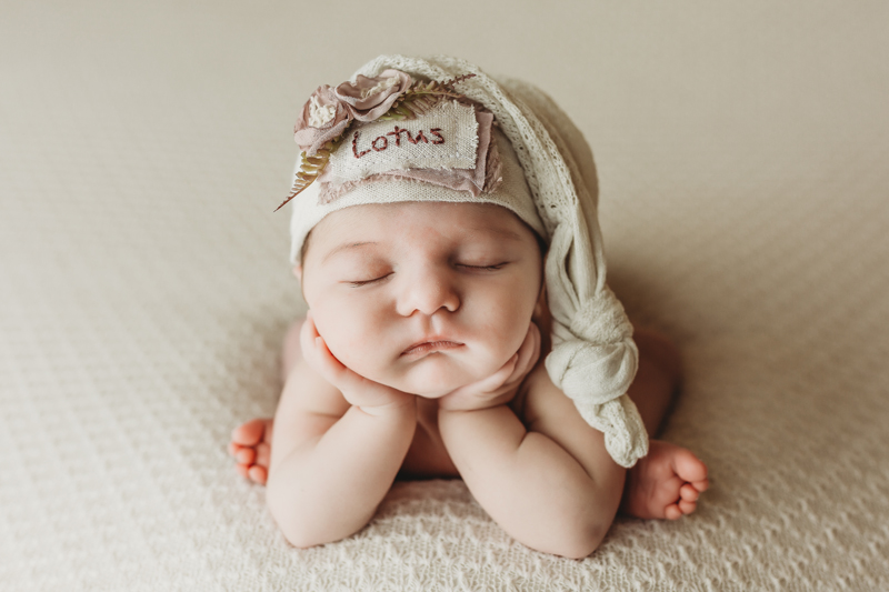 Richmond, Virginia newborn photographer, newborn baby girl in froggy pose on ivory backdrop with off white sleepy cap with the name Lotus on it