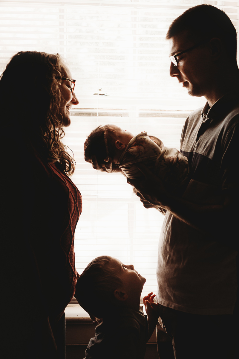 Richmond, Virginia newborn photographer dad holding baby girl in front of a window with mom looking down at baby and big brother looking up