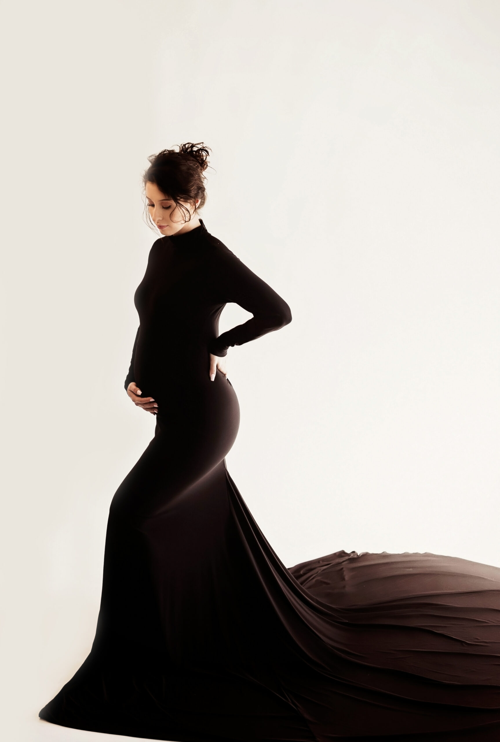 Wichita Falls, Texas maternity photographer, Wichita Falls, Texas newborn photographer, white woman in black gown standing on white backdrop with hand on pregnant belly