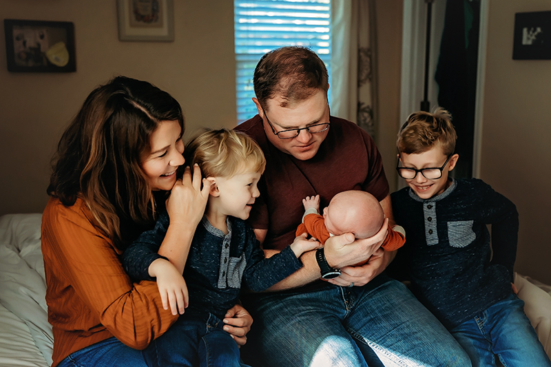 newborn photography in Richmond, Virginia with family of five with dad holding newborn boy and brothers and mom sitting on side of the bed looking at baby