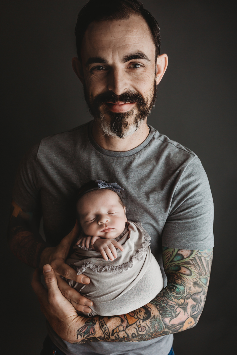Richmond, Virginia newborn photographer, dad with beard in gray shirt holding newborn baby girl wrapped in gray ruffled fabric smiling and looking at camera