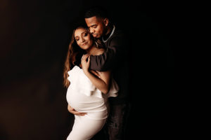 Henrico photographer, pregnant woman in white dress cradling her baby bump with husband holding her shoulder from behind
