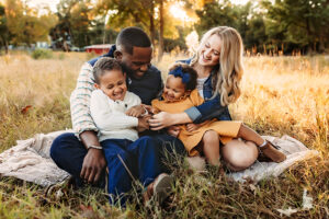 Moseley, Virginia photographer, family of four with black dad and white mom snuggling and laughing with their children sitting on the ground in a field